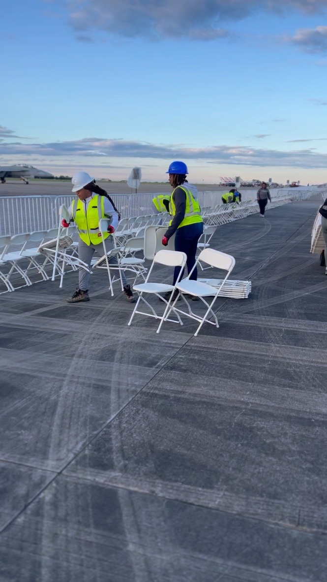 Two crew members carrying white folding chairs during an outdoor event setup