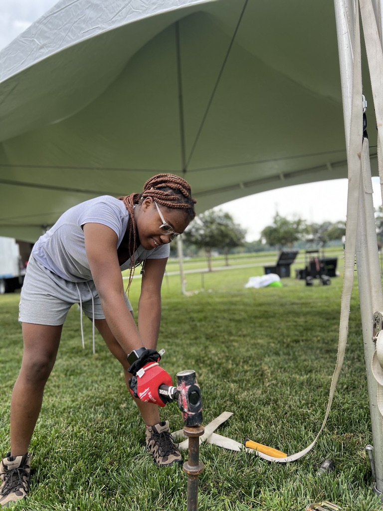 Crew member wearing gloves and safety glasses driving a tent stake into the ground