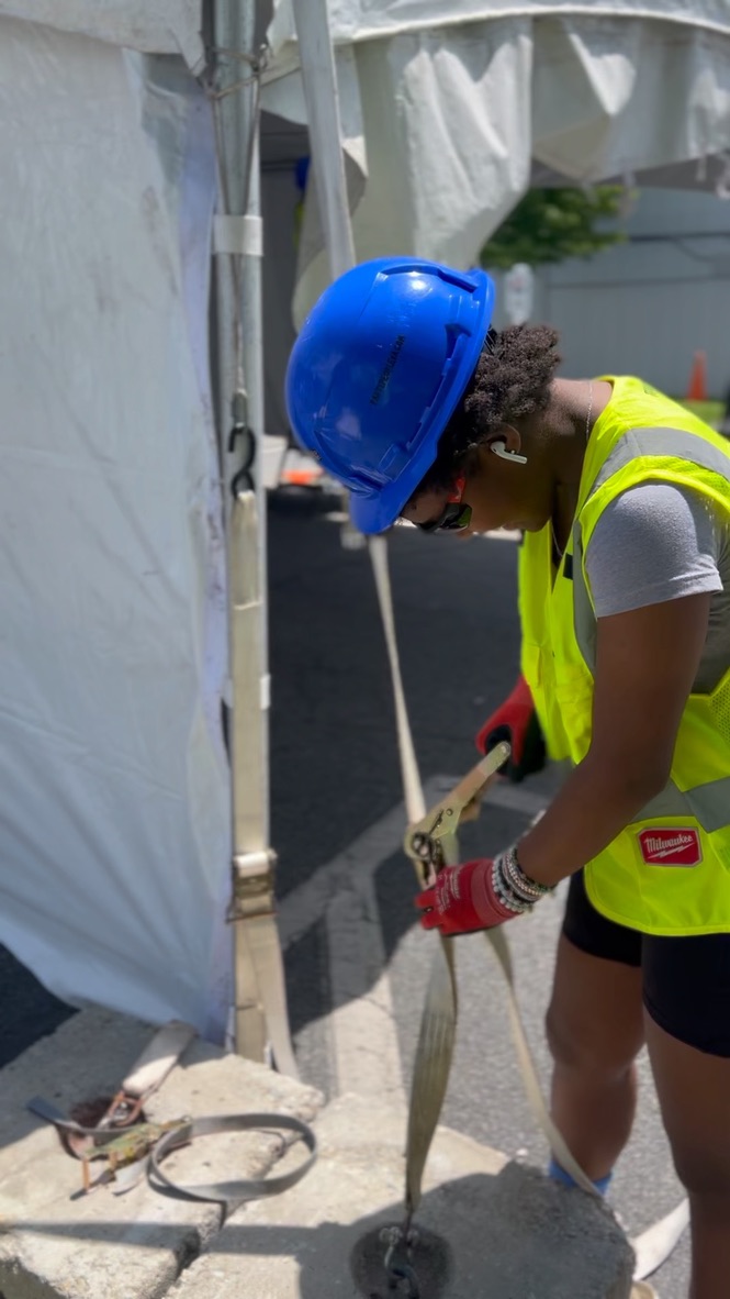 Crew member in a blue hard hat and safety vest tightening an event tent strap